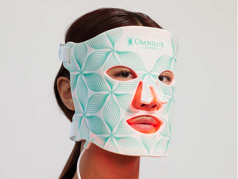 A person wearing an Omnilux Contour light therapy mask with teal geometric patterns and cutouts for the eyes, nose, and mouth. The mask covers the entire face and is secured with side straps. Fearrington Village