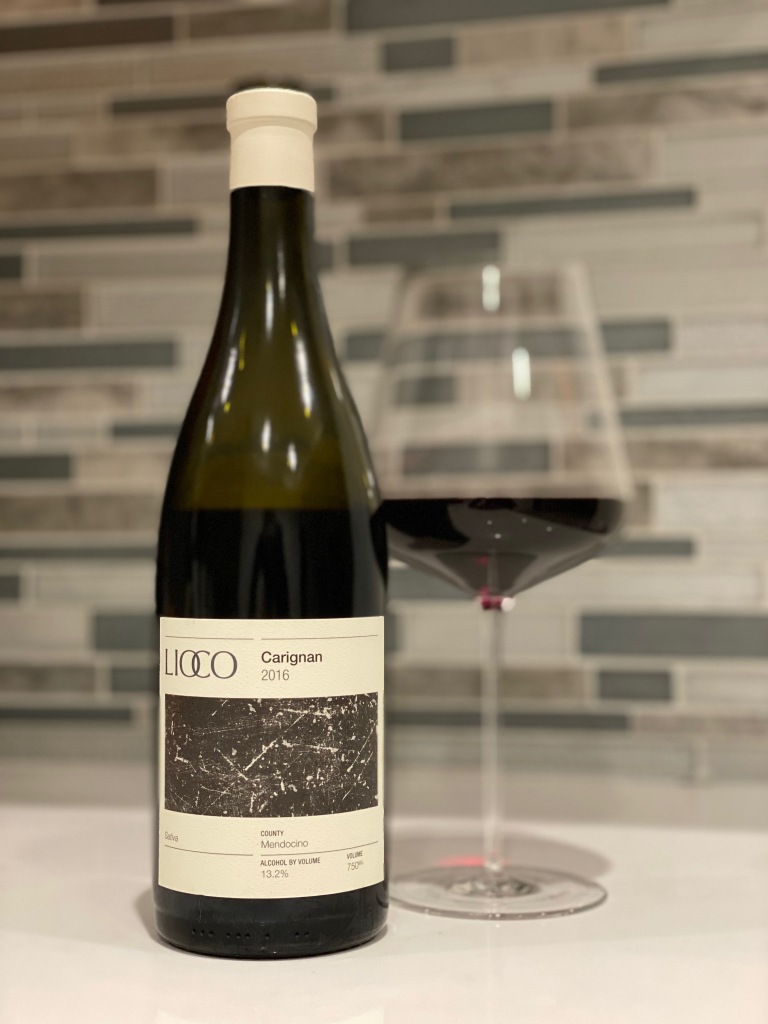 A bottle of Lioco Carignan 2016 red wine stands next to a filled wine glass on a counter, with a modern tiled backsplash in the background. Fearrington Village