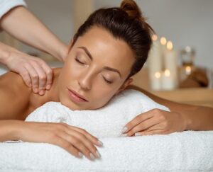 A woman with closed eyes relaxes on a massage table, resting her head on a white towel, while receiving a shoulder massage. Soft lighting and blurred candles create a calming spa atmosphere in the background. Fearrington Village
