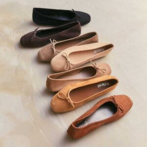 Six pairs of ballet flats in a gradient of neutral colors, from black at the top to light brown and tan shades, to rust at the bottom, arranged in a straight vertical line on a light surface. Fearrington Village