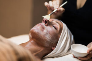 A person with a headband lies on a spa table with eyes closed as a facial mask is applied to their face using a brush. An aesthetician holds a small bowl in one hand and applies the mask with the other. Fearrington Village