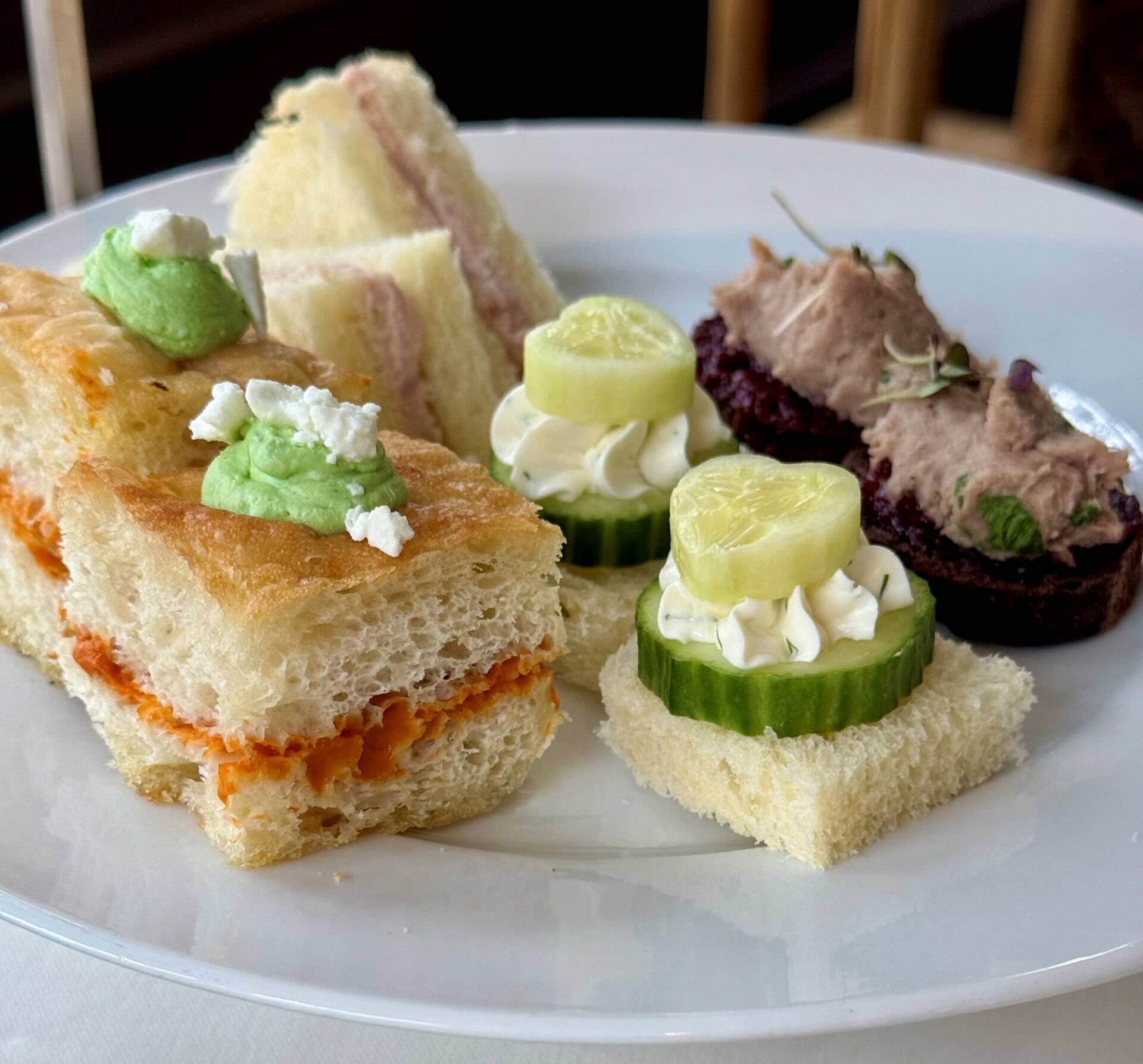 A white plate with assorted tea sandwiches, including ham sandwiches, focaccia with spread, cucumber and cream cheese on bread squares, and a small toast with a dark pâté and garnish. Fearrington Village