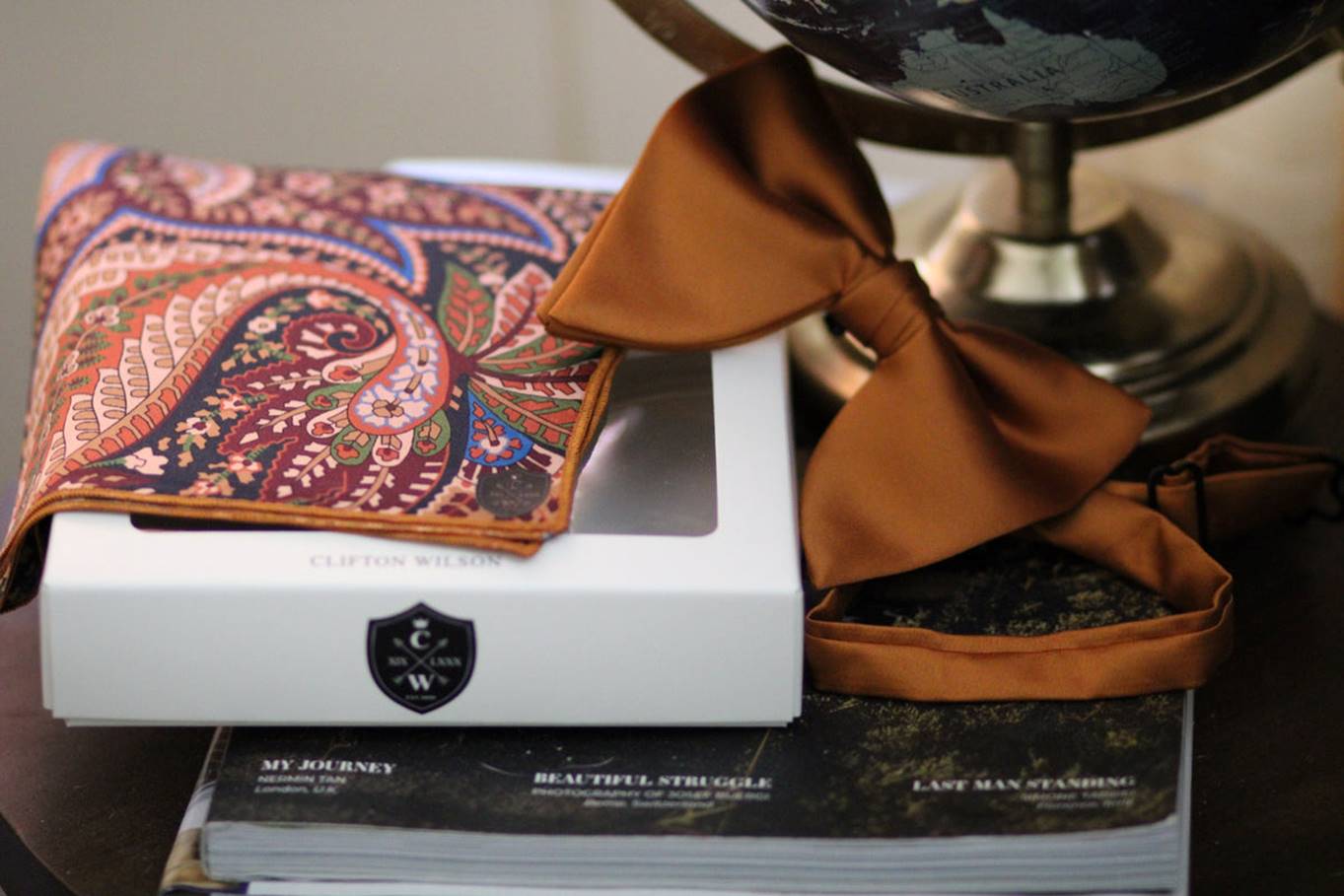 A patterned pocket square, a rust-colored bow tie, and its matching band rest on a white box next to a globe and magazines on a dark wooden surface. Fearrington Village