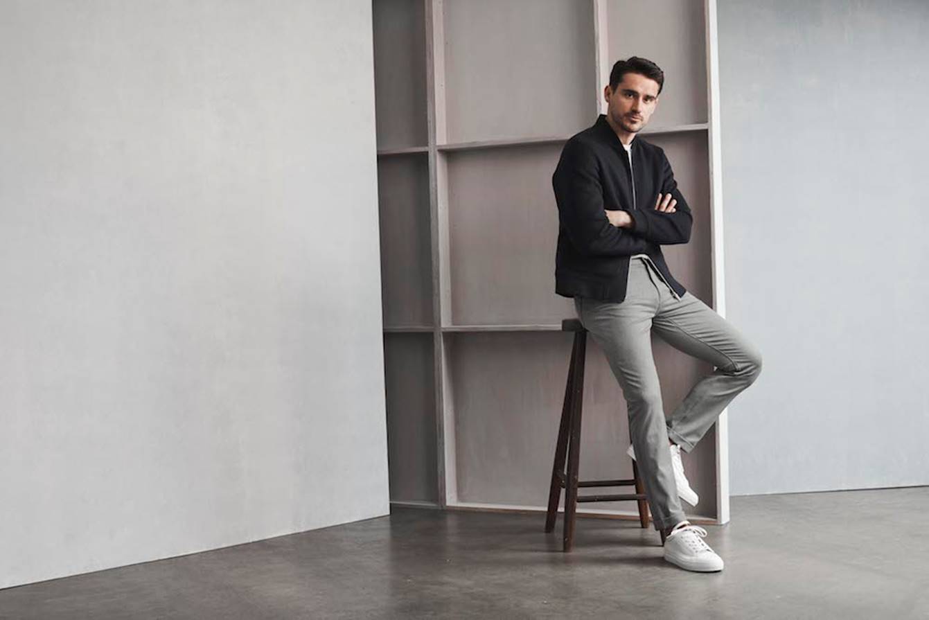 A man in a black jacket, dark shirt, gray pants, and white sneakers sits on a wooden stool in a minimalist room with gray walls and a large empty shelving unit behind him. Fearrington Village