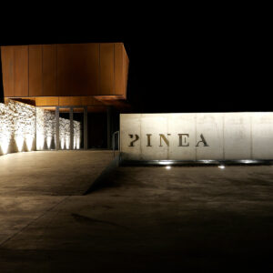 A modern building at night with a stone wall on the left and a large illuminated concrete wall on the right displaying the word "PINEA." The entrance features a metallic, rust-colored structure above glass doors. Fearrington Village