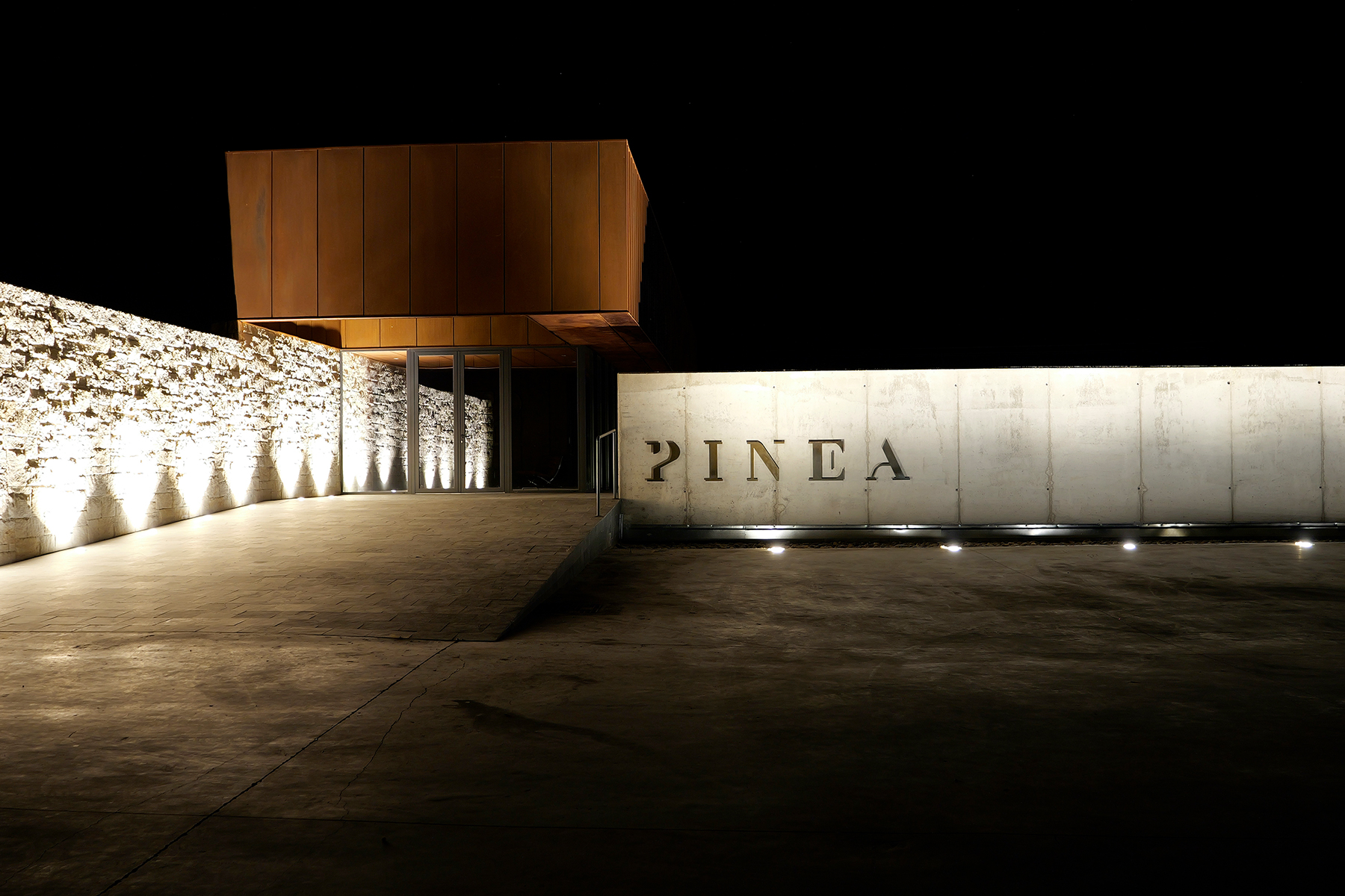 A modern building at night with a stone wall on the left and a large illuminated concrete wall on the right displaying the word "PINEA." The entrance features a metallic, rust-colored structure above glass doors. Fearrington Village