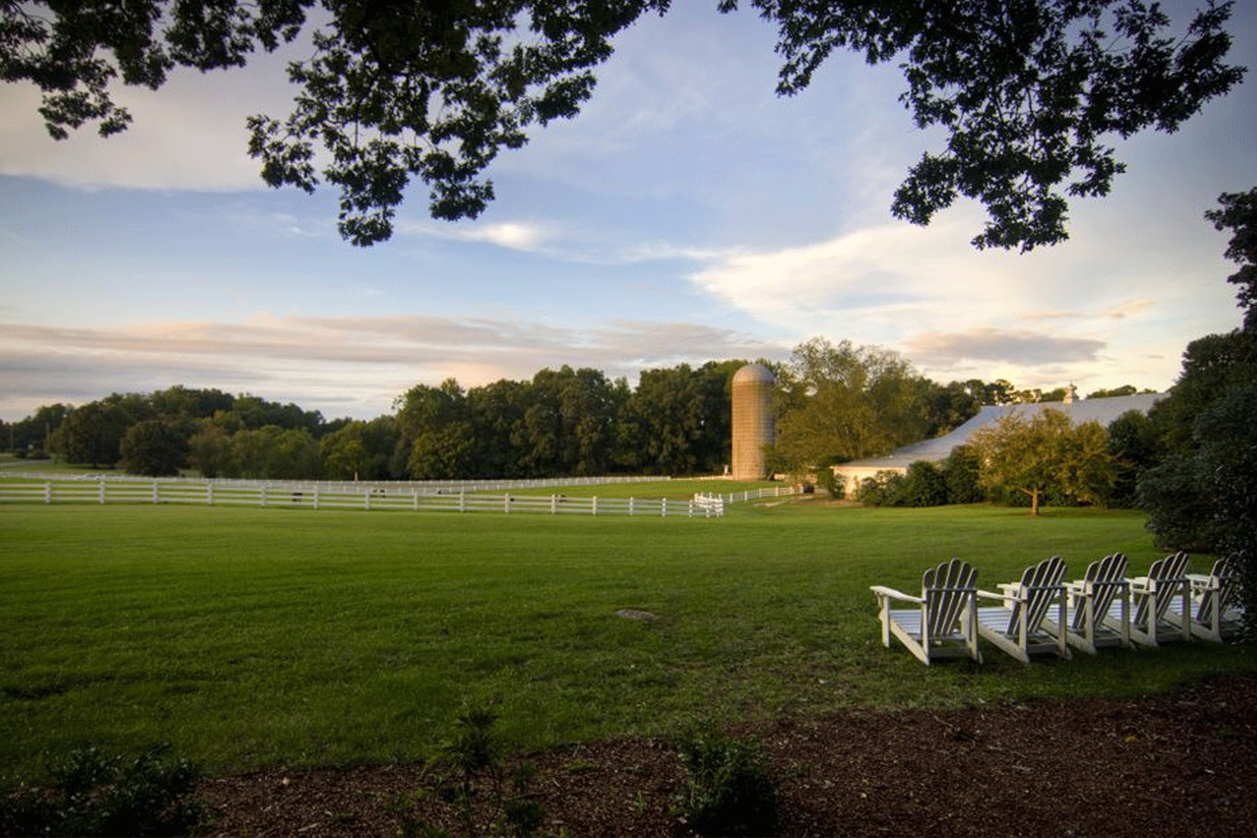 A grassy field with white Adirondack chairs faces a fenced pasture, trees, a silo, and a barn under a partly cloudy sky. The scene is peaceful and rural, framed by overhanging tree branches. Fearrington Village