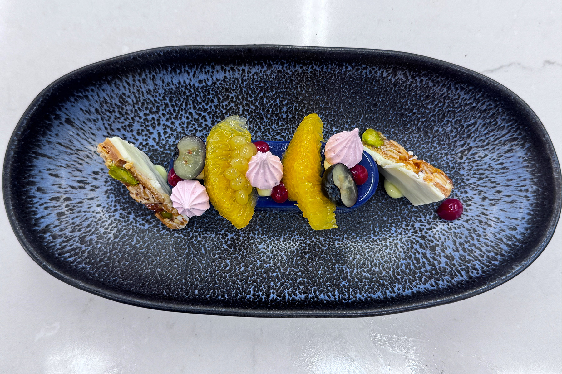 A dark oval plate with artistic dessert arrangement featuring orange segments, pink meringue drops, grapes, red berries, green pistachios, and layered nut crisps on a speckled blue-black background. Fearrington Village