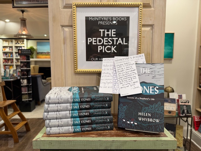 A display in a bookstore features stacked copies of "The Salt Stones" by Helen Whybrow, with one book upright and note cards attached. A framed sign above reads, "The Pedestal Pick" at McIntyre's Books. Fearrington Village