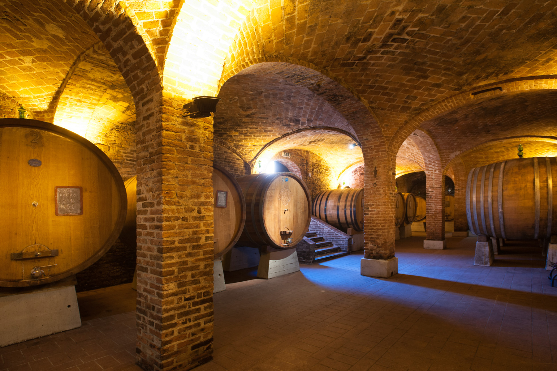 A dimly lit wine cellar with arched brick ceilings and walls, featuring large wooden barrels lined up along the stone floor and ambient yellow lighting creating a warm atmosphere. Fearrington Village