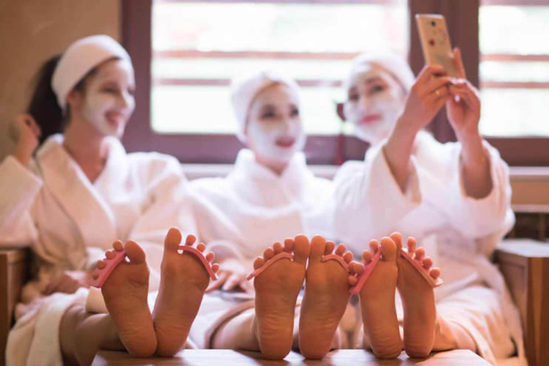Three women in white robes and face masks sit together, smiling and taking a selfie, with their feet in the foreground and toe separators on their toes, enjoying a relaxing spa day. Fearrington Village
