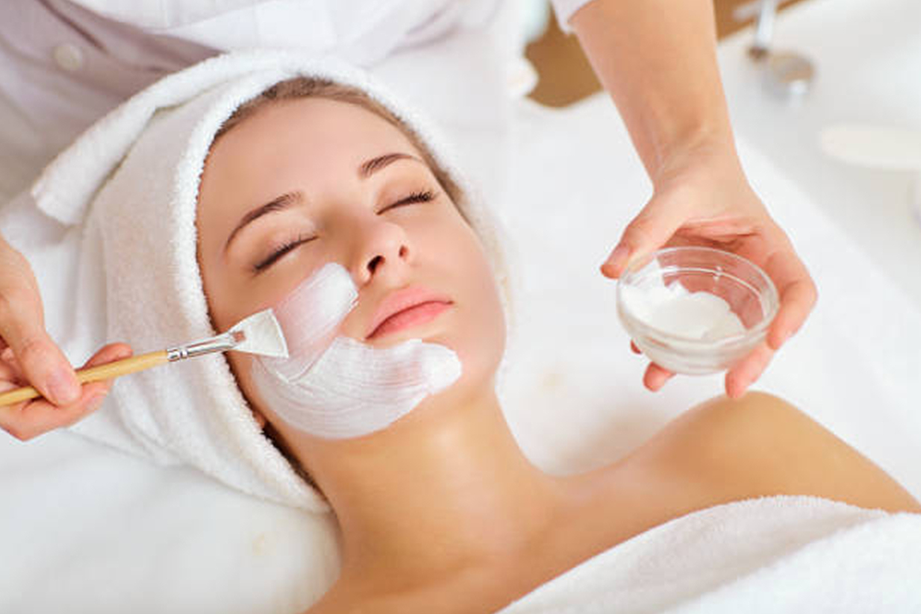 A woman with a towel wrapped around her head receives a facial treatment as a beautician applies a white face mask using a brush. She is lying down with her eyes closed, looking relaxed. Fearrington Village
