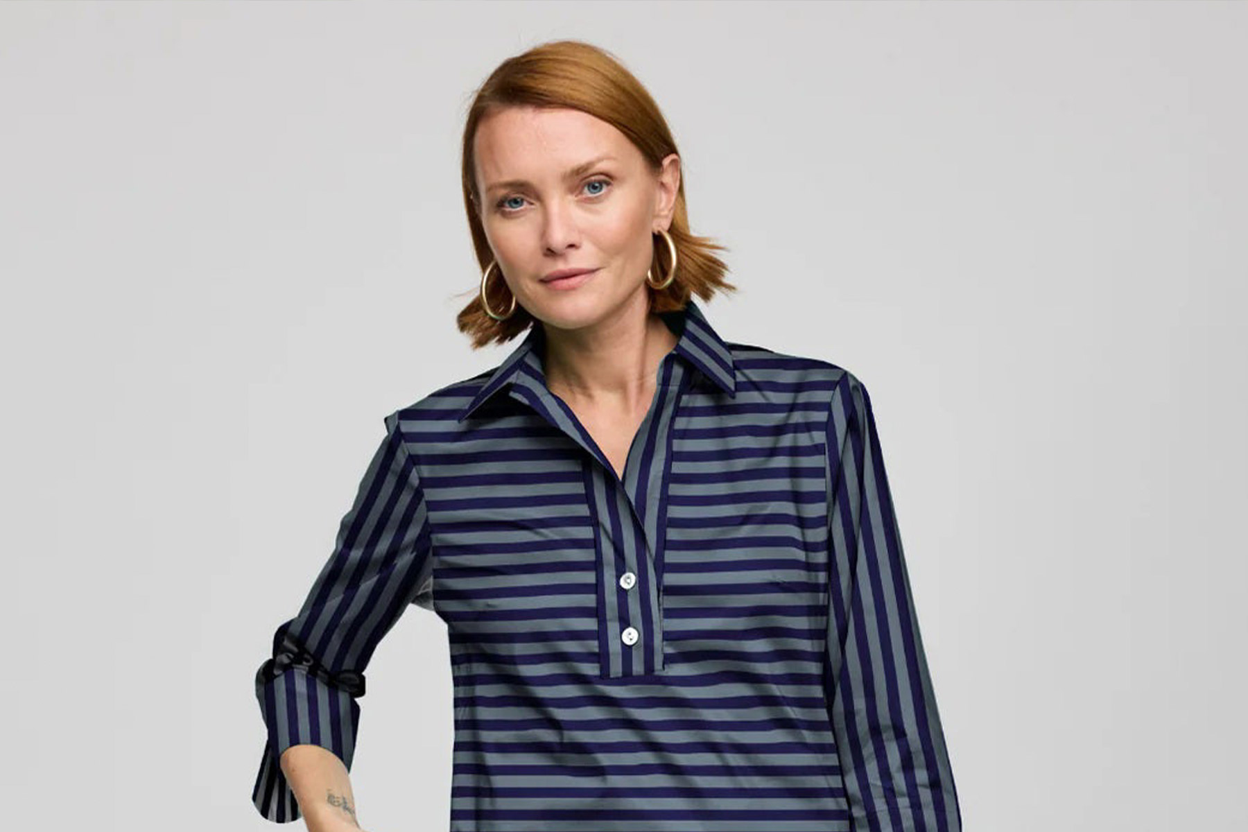 Woman with short red hair wearing large hoop earrings and a navy blue collared shirt with horizontal and vertical stripes, standing against a plain light background. Fearrington Village