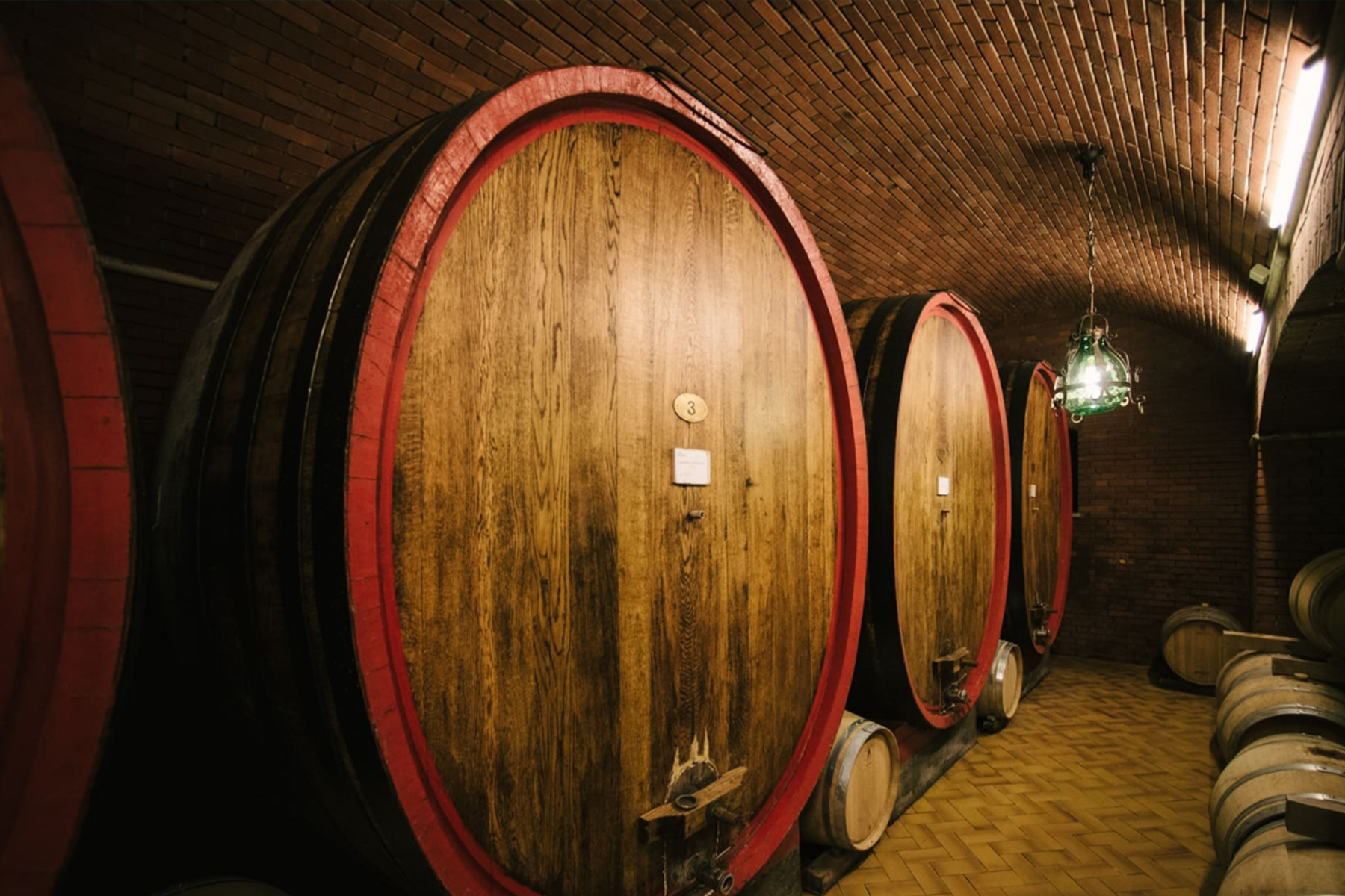 Large wooden wine barrels with red rims are lined up against a brick wall in a dimly lit cellar, with a hanging green lamp providing light and smaller barrels visible in the background. Fearrington Village