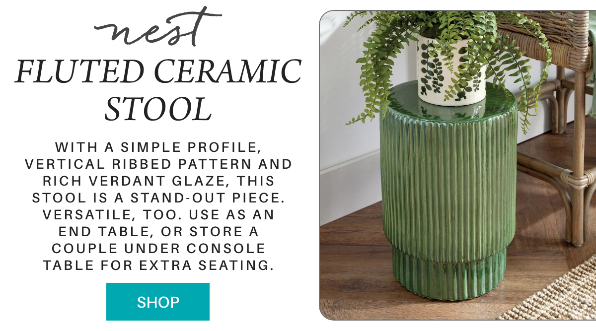 Green fluted ceramic stool with vertical ribbed design, used as a small table holding a potted fern next to a wicker chair on a wood floor. Text describes its versatility as seating or a table. A blue “shop” button is visible. "With a simple profile, vertical ribbed pattern and rich verdant glaze, this stool is a stand-out piece. Versatile, too. Use as an end table, or store a couple under console table for extra seating." Fearrington Village