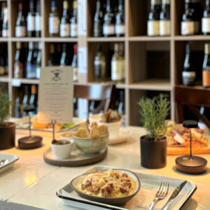 A table set with plated food, cutlery, small potted herbs, and wine glasses sits in front of shelves filled with various bottles of wine in a cozy restaurant setting. Fearrington Village