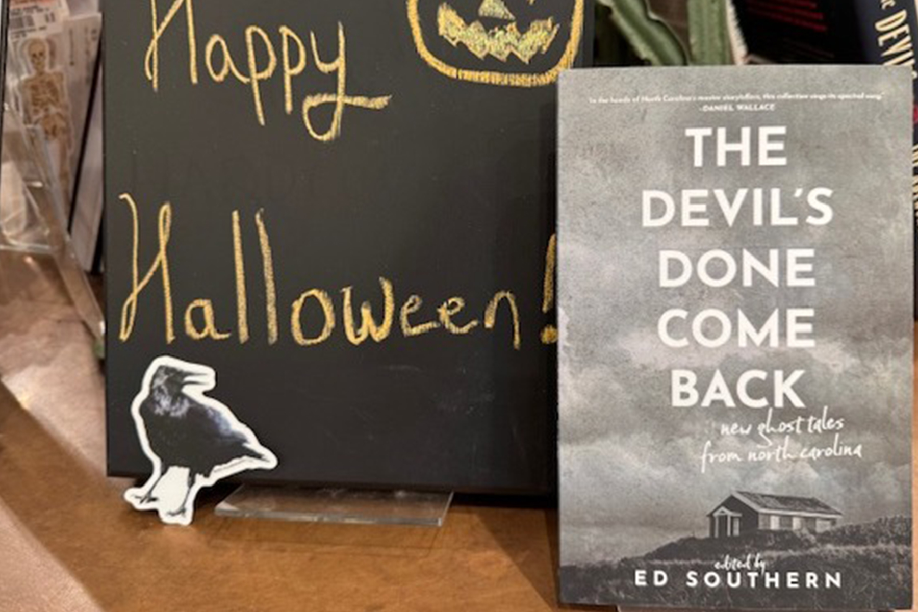 A book titled "The Devil’s Done Come Back" stands upright next to a chalkboard sign that says “Happy Halloween!” with a pumpkin drawing, and a small raven cutout in front of the sign. Fearrington Village
