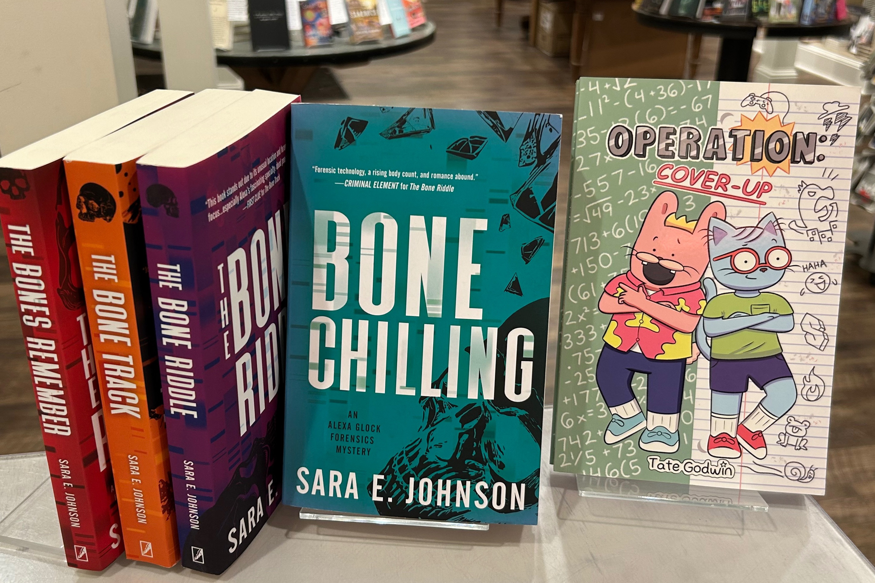 Five books are displayed upright on a table: four Sara E. Johnson mystery novels with bold, colorful covers, and a children’s book titled "Operation: Cover-Up" featuring illustrated animal characters and math doodles. Fearrington Village