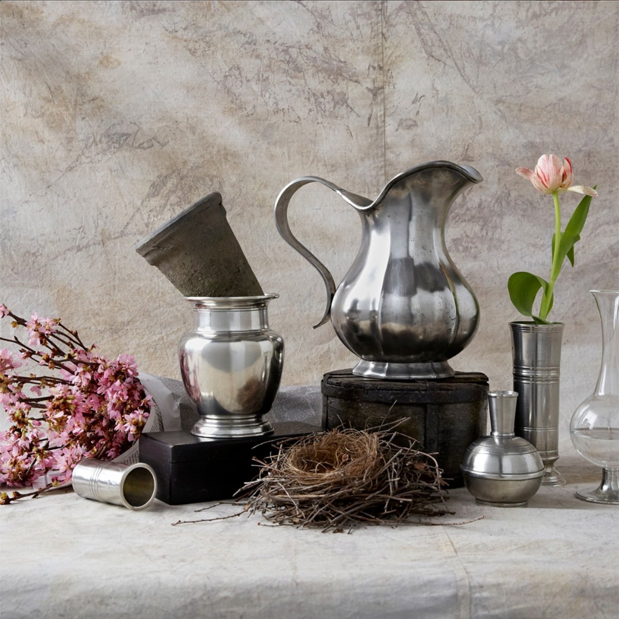 A still life arrangement with silver vessels, a nest, pink blossoms, a single pink tulip in a vase, and a pale textured background. The scene blends natural and metallic elements on a soft, draped surface. Fearrington Village
