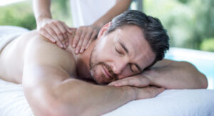 The Spa Edit - Swedish vs. Deep Tissue Massage: Which Is Right for You? 3 A man with short brown hair and a beard lies face down on a massage table with his eyes closed, smiling while receiving a shoulder massage from a person whose hands are visible. Fearrington Village