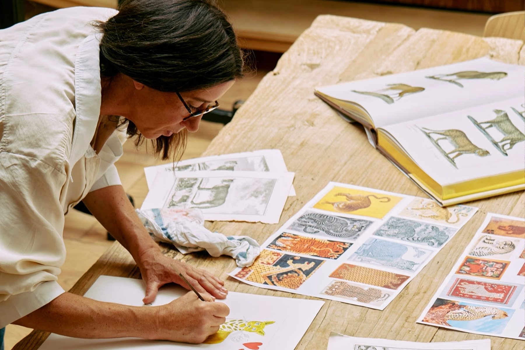 A person wearing glasses draws an animal illustration at a wooden table covered with art prints and an open book displaying animal drawings. Fearrington Village