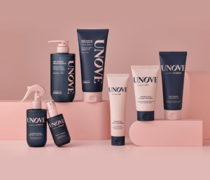 A set of UNOVE hair care products, including bottles, tubes, and a spray, displayed on pink platforms against a pink background. The packaging is mainly dark blue with white text and a few light pink accents. Fearrington Village