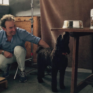 A man wearing a blue shirt and white pants crouches and smiles while petting a curly black dog next to a table with a metal dog bowl and glass jars. The setting appears to be indoors with soft, natural light. Fearrington Village