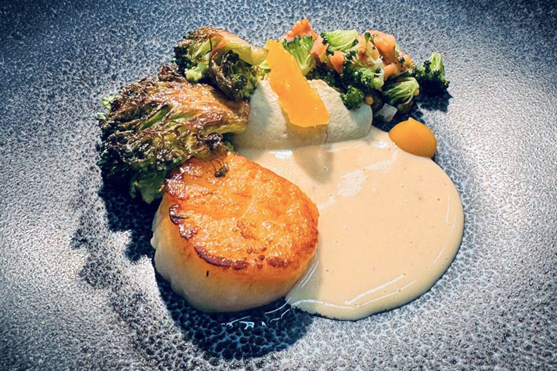 A seared scallop is plated with creamy sauce, roasted broccoli, a dollop of yellow puree, diced vegetables, and a citrus segment on a textured dark plate. The presentation is colorful and artistic. Fearrington Village