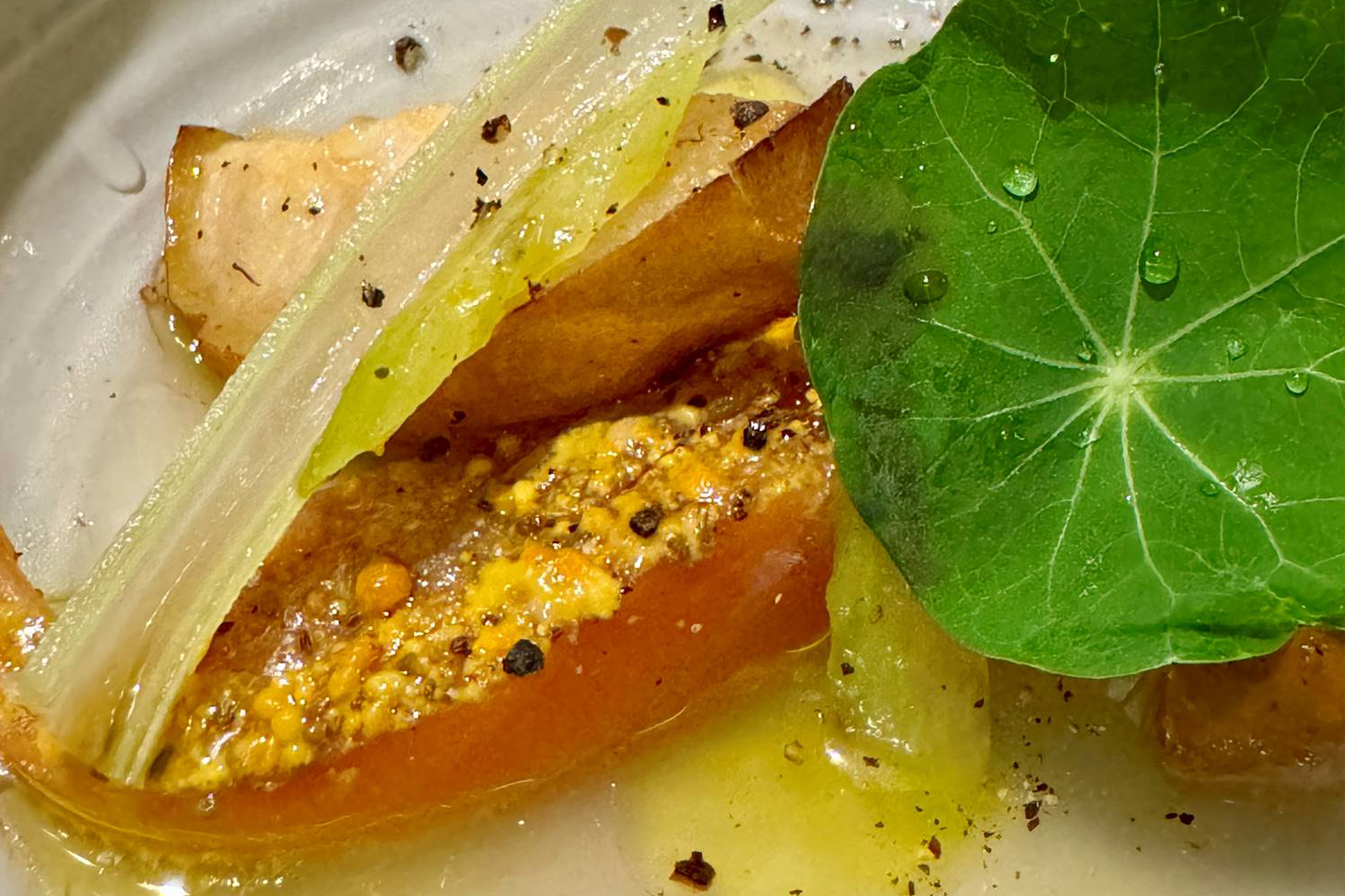 A close-up of a plated gourmet dish featuring a large green leaf, a slice of tomato topped with spices and oil, celery stalks, and roasted potato pieces, all garnished with cracked black pepper. Fearrington Village