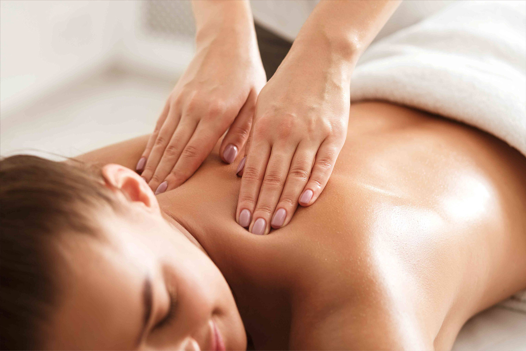 A person receiving a relaxing back massage from a massage therapist, with gentle hands pressing on their upper back. The person is lying face down on a massage table, covered by a white towel. Fearrington Village
