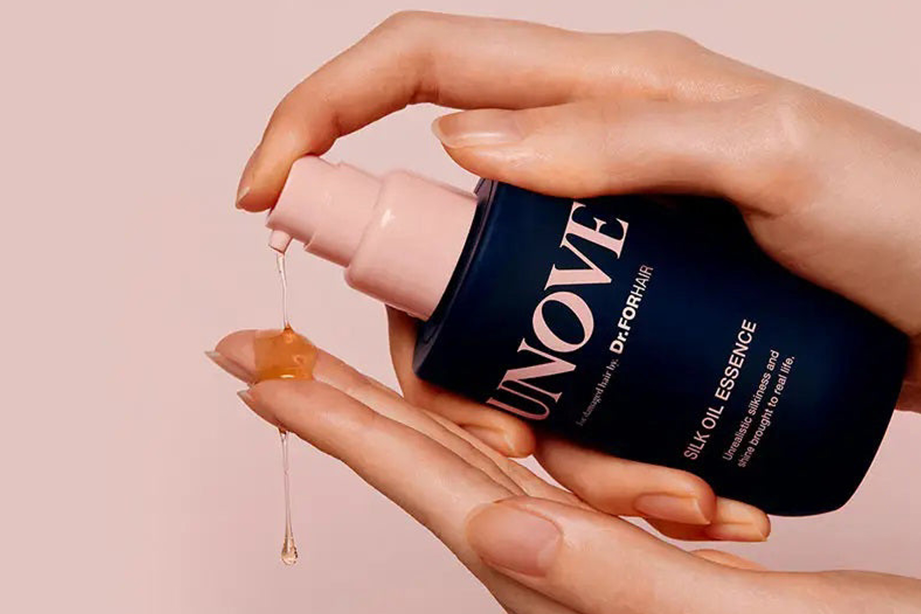 A person dispenses a clear, golden liquid from a blue and pink UNOVE Silk Oil Essence bottle onto their palm against a light pink background. Fearrington Village