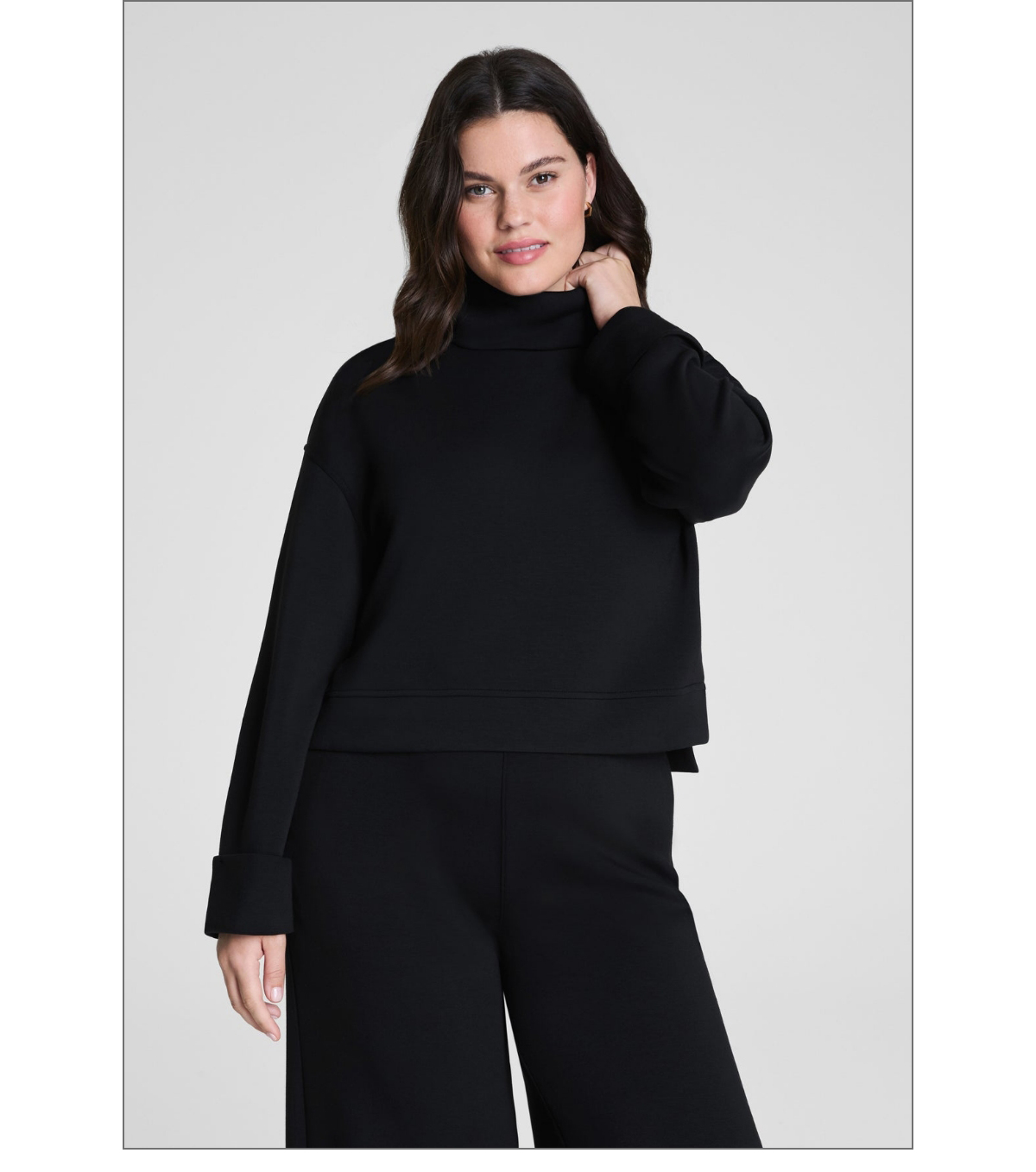 A woman with wavy dark hair is wearing a loose black turtleneck sweater with wide sleeves and matching black pants. She is standing against a plain light background, looking at the camera with a slight smile. Fearrington Village