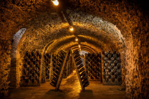 Dimly lit wine cellar with arched stone ceilings, walls, and floors, lined with racks holding many bottles of wine at an angle; yellow lights create a warm, rustic atmosphere. Fearrington Village