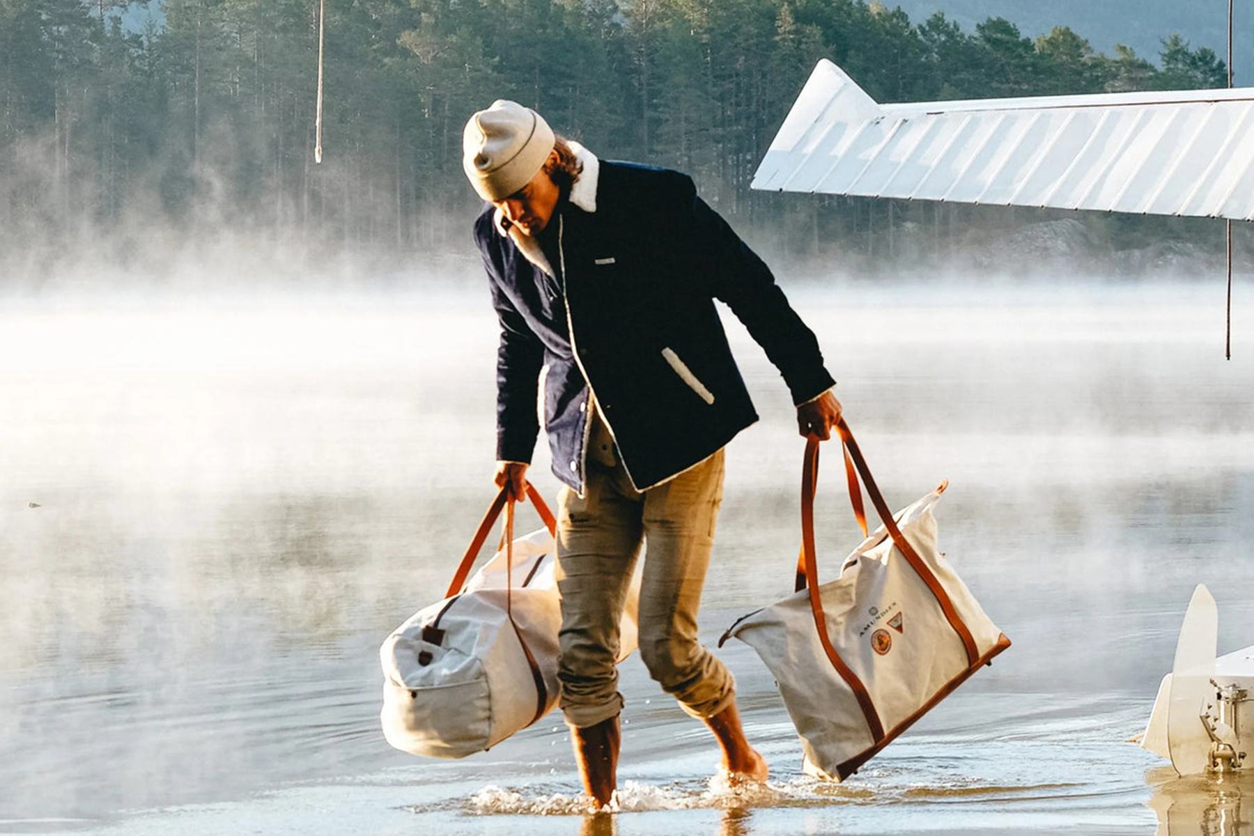 A person wearing a white beanie, navy jacket, and rolled-up pants walks barefoot through shallow water, carrying two large duffel bags. Mist rises from the water and a seaplane wing is visible in the background. Fearrington Village