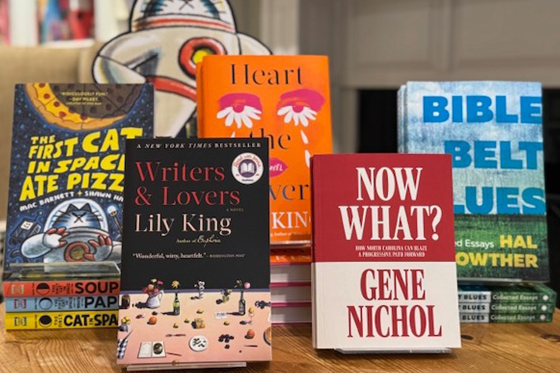 A selection of books displayed upright on a table, including "Writers & Lovers," "The First Cat in Space Ate Pizza," "Soup of the Day," "Heart," "Now What?," and "Bible Belt Blues. Fearrington Village
