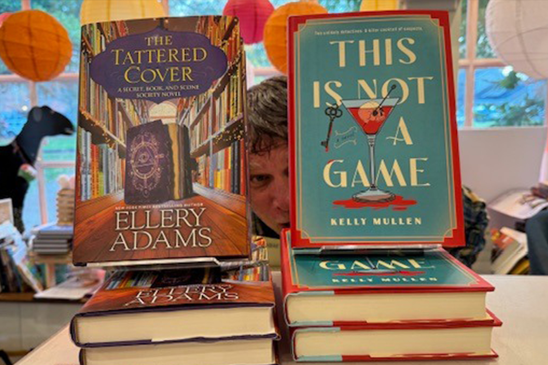 A person peeks between two stacks of books titled "The Tattered Cover" by Ellery Adams and "This Is Not a Game" by Kelly Mullen in a brightly lit bookstore. Colorful paper lanterns hang in the background. Fearrington Village