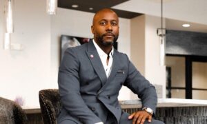 A man in a gray suit sits confidently on a modern chair at a counter, looking directly at the camera in a stylish, contemporary indoor setting. Fearrington Village