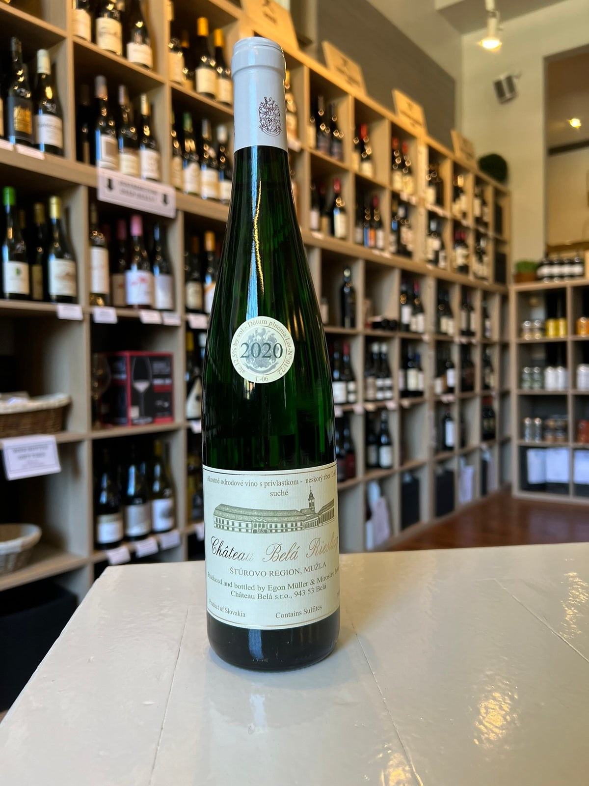 A green bottle of Château Bella Tétény wine stands on a white counter in a wine shop, surrounded by shelves filled with various other wine bottles. The label indicates the vintage year as 2020. Fearrington Village