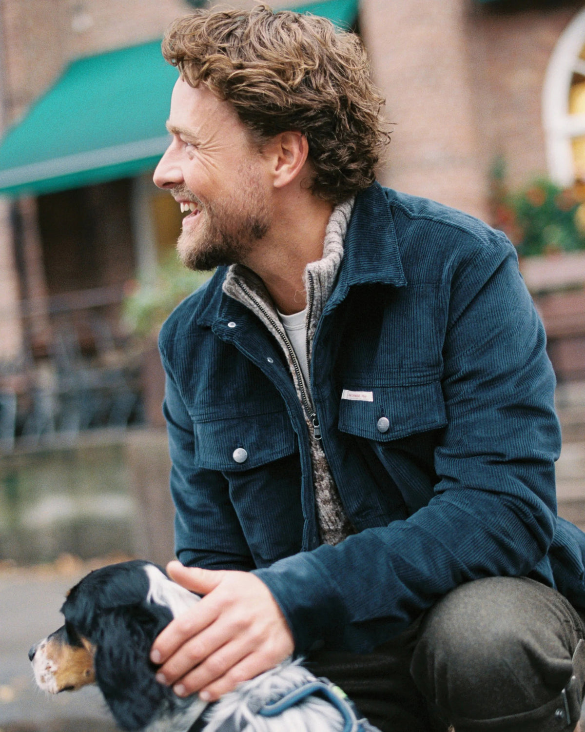 A man with curly hair and a beard, wearing a blue jacket, smiles while sitting outdoors and petting a black and white dog. The background features a building with a green awning. Fearrington Village