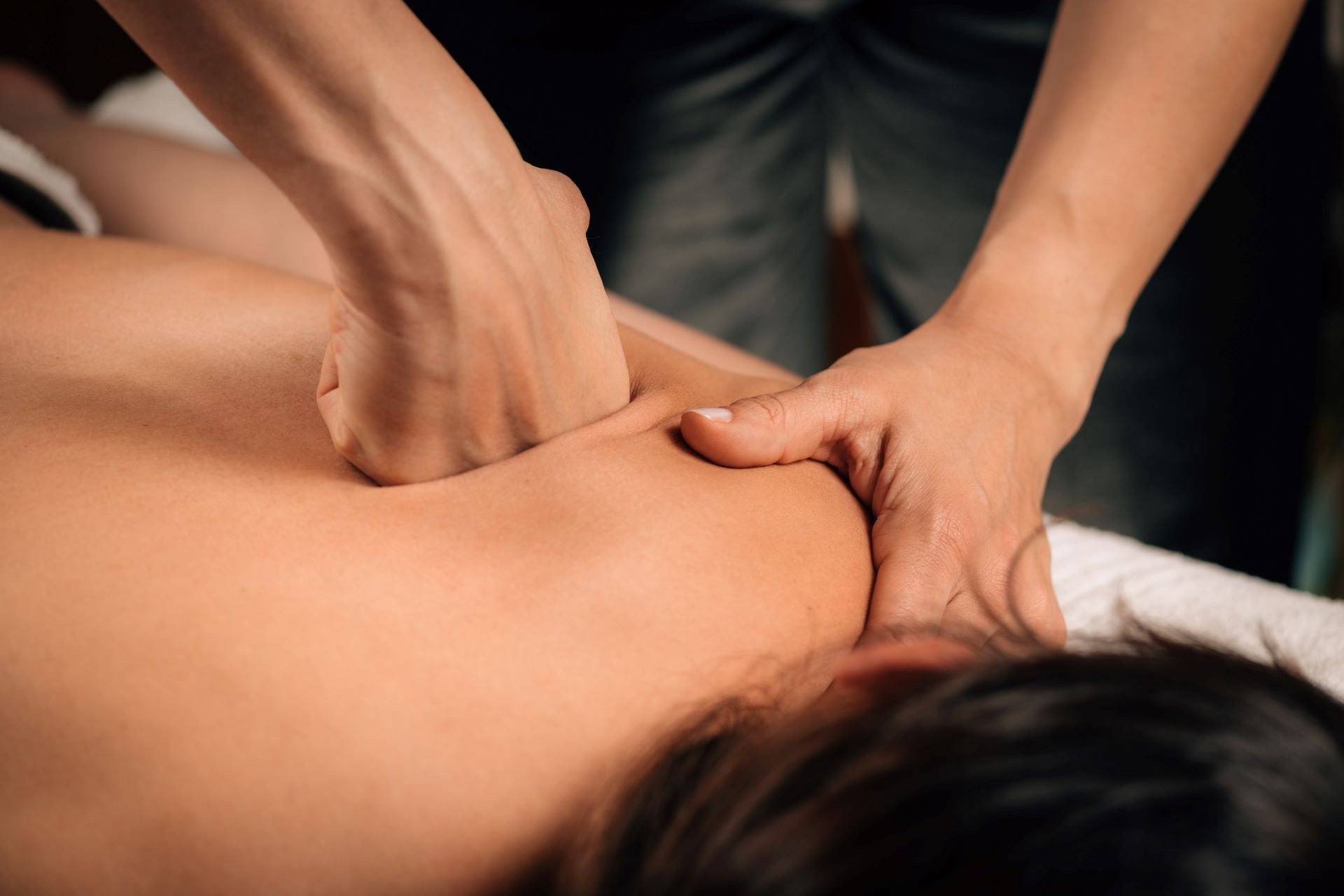 A close-up of a person receiving a deep tissue massage, showing hands pressing firmly into the muscles of their upper back and shoulder area. The person is lying face down on a massage table. Fearrington Village