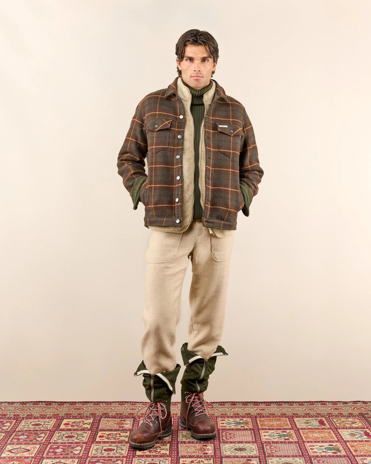 A man stands on a patterned rug against a plain background, wearing a brown plaid jacket over a green turtleneck, beige pants tucked into green gaiters, and brown lace-up boots. His hands are in his jacket pockets. Fearrington Village