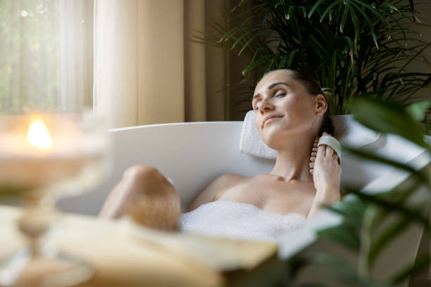The Spa Edit - Relaxation Techniques for Busy Minds This Fall 3 A woman relaxes in a bubble-filled bathtub, leaning back with a towel behind her head and a serene expression, surrounded by green plants and a lit candle in the foreground. Fearrington Village