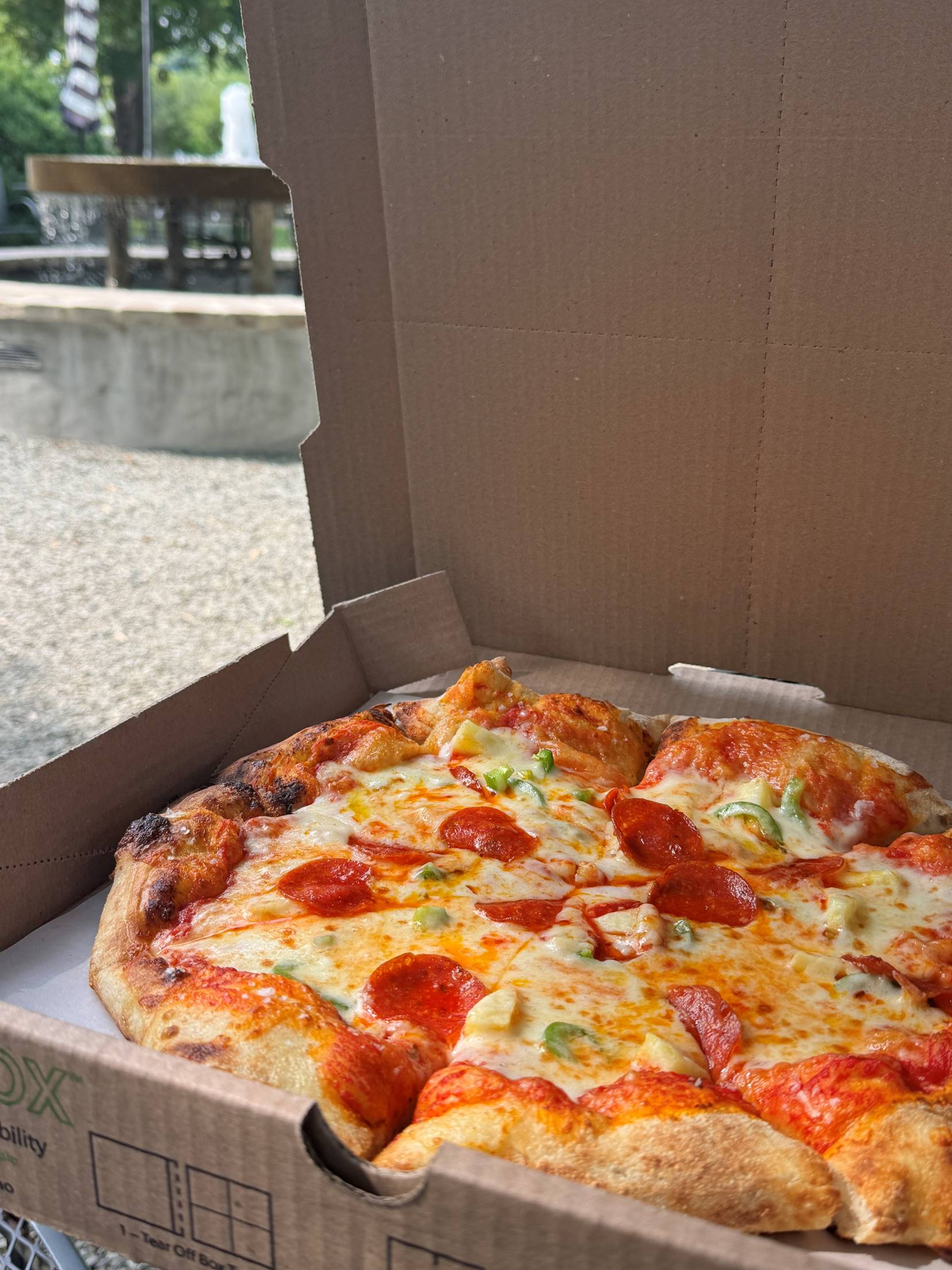A pizza with pepperoni and green peppers sits in an open cardboard box outdoors, with a picnic table and trees visible in the blurred background. Fearrington Village