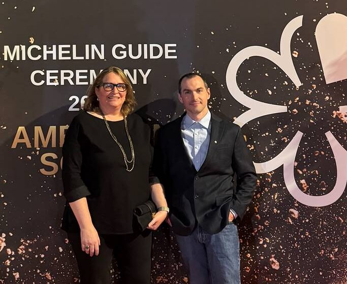 Two people in formal attire stand in front of a black and gold backdrop at the Michelin Guide Ceremony 2023, featuring a large Michelin star logo and celebratory design elements. Fearrington Village