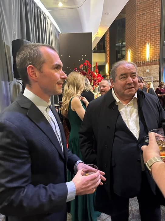 Two men in suits stand at a formal indoor event, holding drinks and conversing. In the background, other guests mingle near a floral arrangement and draped curtains, with exposed brick walls visible. Fearrington Village