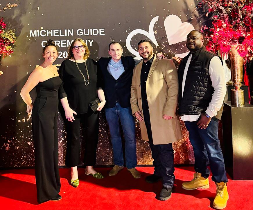 Five people stand smiling on a red carpet at the Michelin Guide Ceremony, 2024, posed in front of a black backdrop with large white lettering and festive red floral arrangements. Fearrington Village