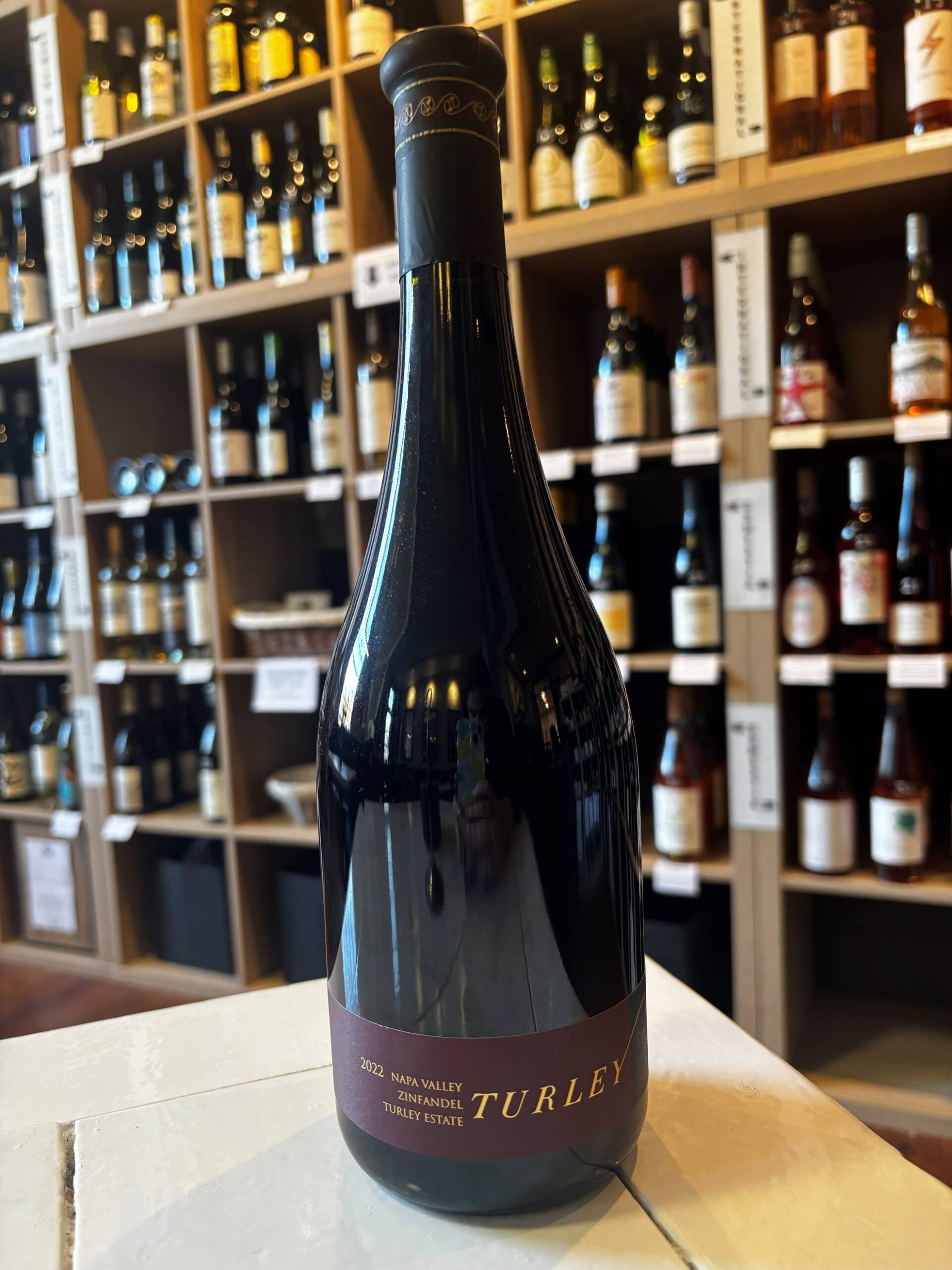 A bottle of 2022 Turley Napa Valley Estate red wine is displayed on a white surface, with shelves of various wine bottles in the background at a wine shop. Fearrington Village
