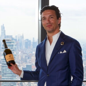 A man in a blue suit stands by a window with a city skyline in the background, smiling while holding a wine bottle with a beige label and black logo. Fearrington Village