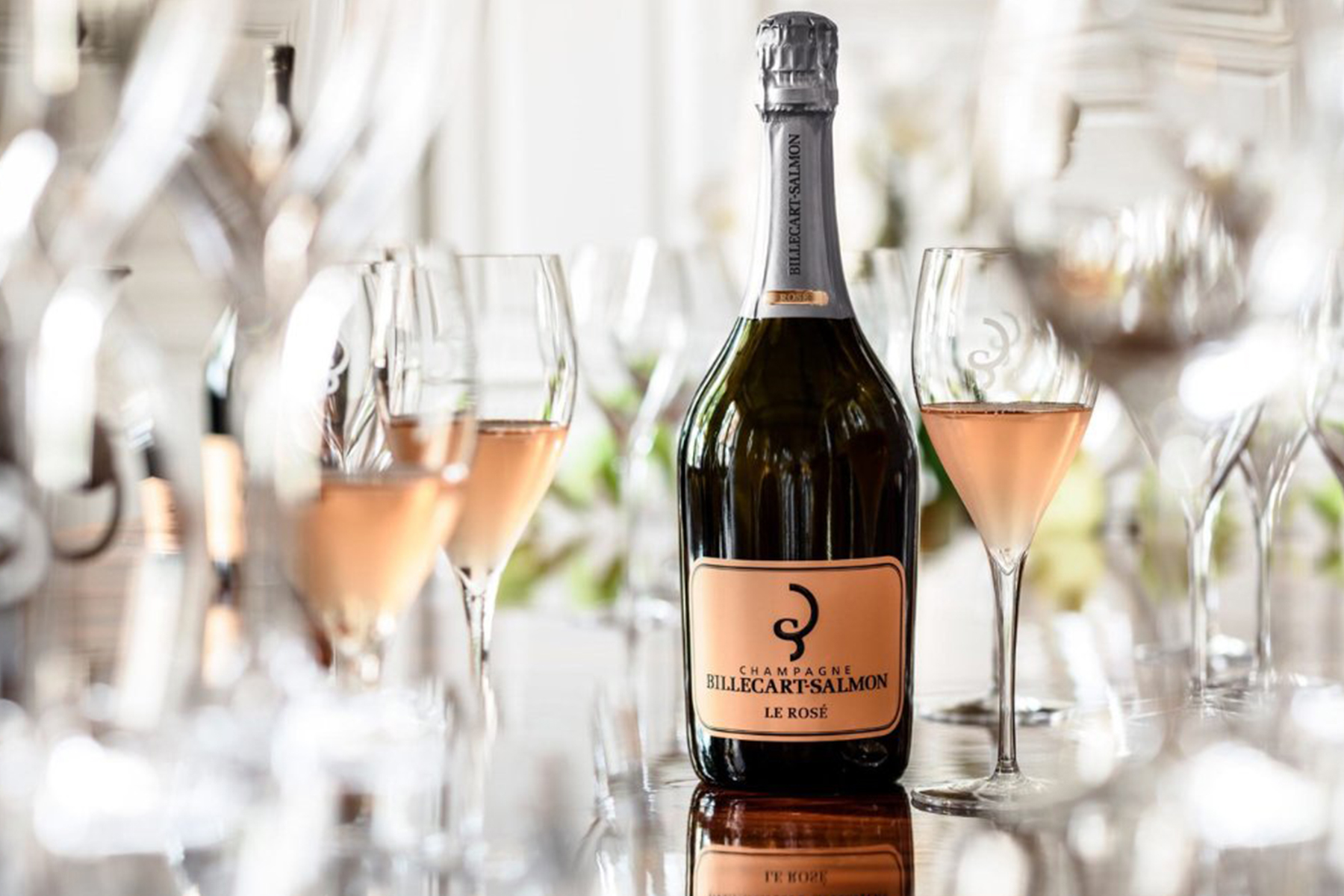 A bottle of Billecart-Salmon Le Rosé champagne is surrounded by several filled and empty wine glasses, with reflections visible on the shiny surface beneath. The scene suggests a celebratory or elegant setting. Fearrington Village