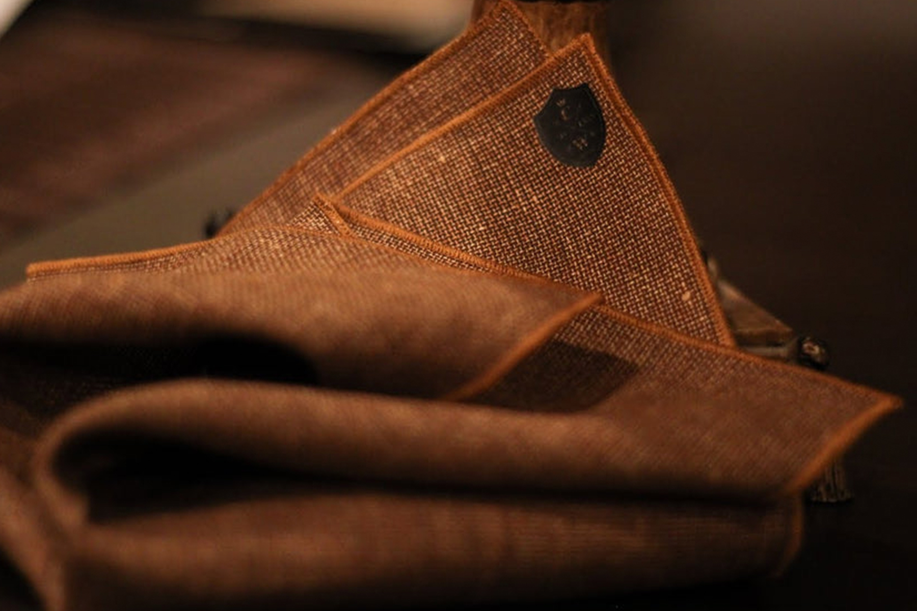 A close-up of neatly folded brown fabric with orange trim, placed on a dark surface. The fabric appears textured and refined, with a small black shield-shaped tag visible in the background. Fearrington Village